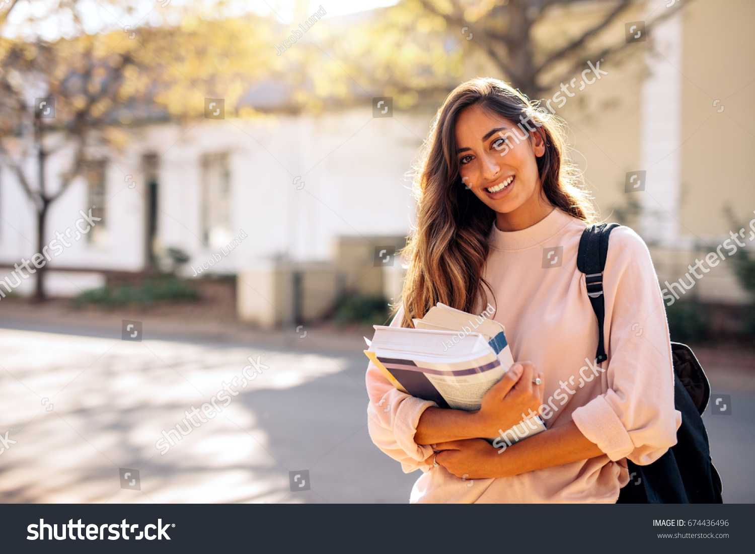 Beautiful young woman with backpack and books outdoors. College student carrying lots of books in college campus. #674436496