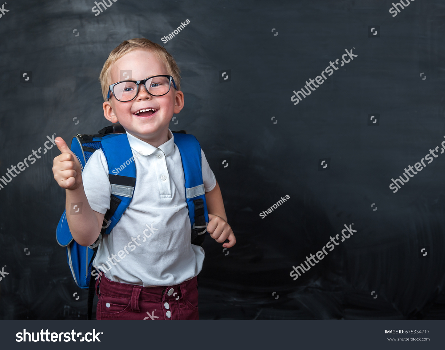 Happy smiling boy in glasses with thumb up is going to school for the first time. Child with school bag and book. Kid indoors of the class room with blackboard on a background. Back to school. #675334717