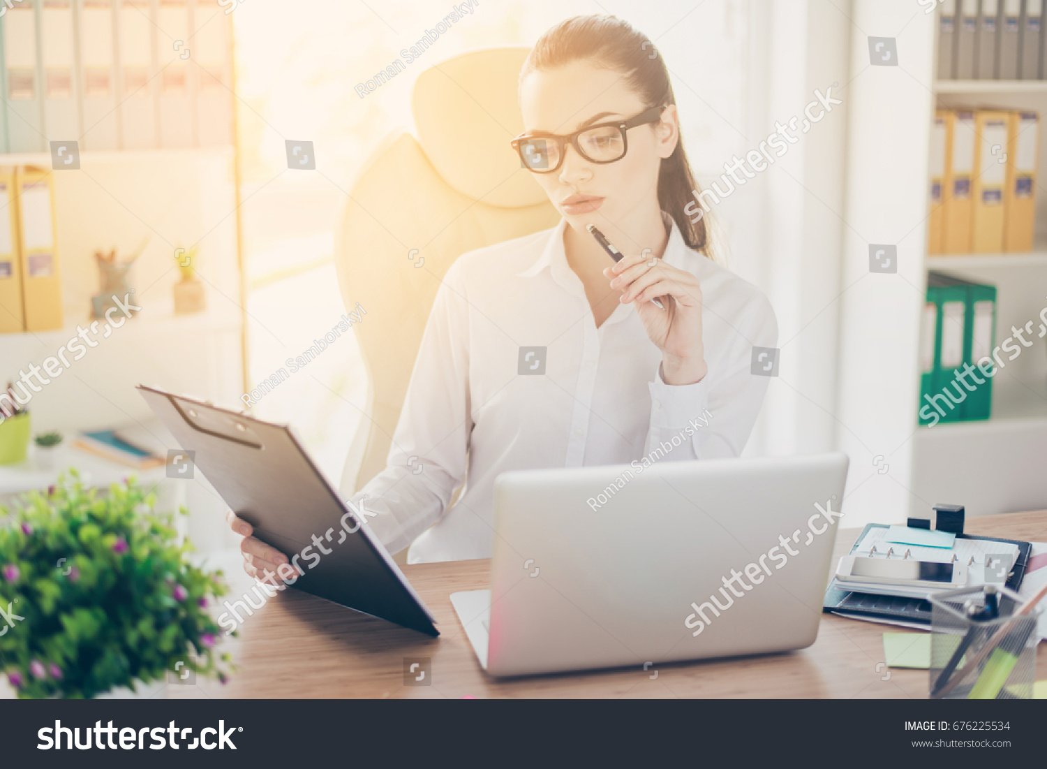 Portrait of serious young businesswoman in formal wear, sitting at her work place and concentrated on data from report in the clip board #676225534