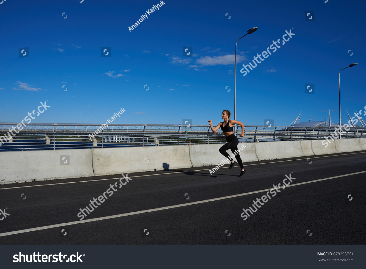 Freeze action shot of attractive determined young female athlete wearing black sportsbra and leggings running in the stadium, preparing for marathon or contest. Endurance and determination concept #678353761