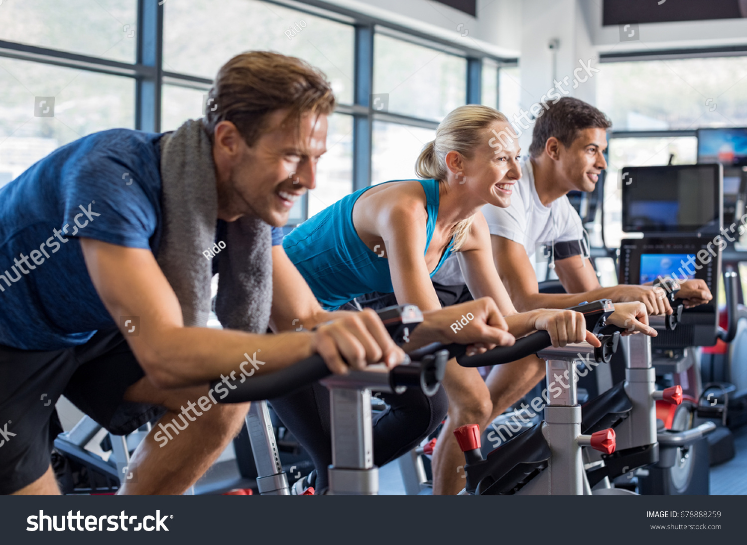 Group of smiling friends at gym exercising on stationary bike. Happy cheerful athletes training on exercise bike. Young men and woman working out at a class in the gym. #678888259