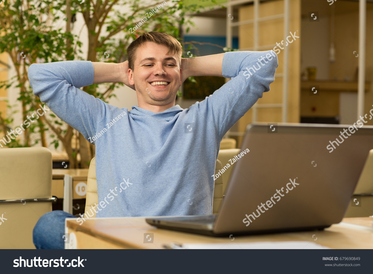 Happy young businessman sitting relaxed at his workplace smiling joyfully after finishing project at the end of the working day technology laptop online positivity confidence success achievement #679690849