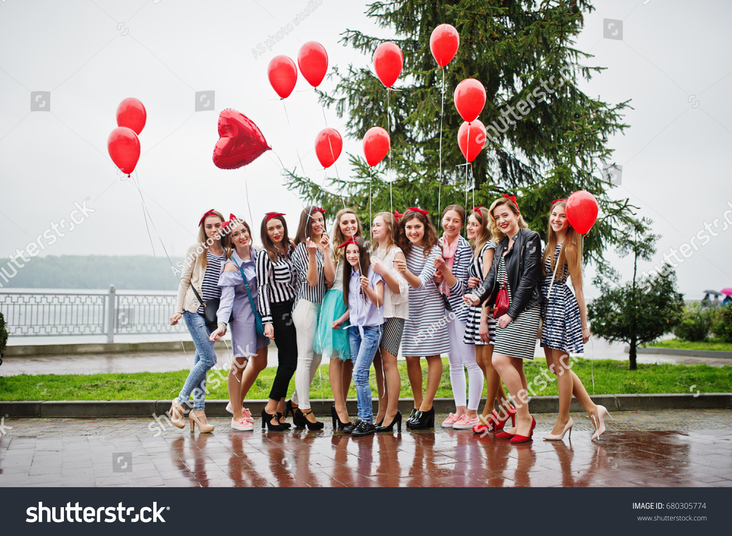 Eleven amazingly-looking braidsmaids with stunning bride posing with red heart-shaped balloons on the pavement against the lake in the background. #680305774