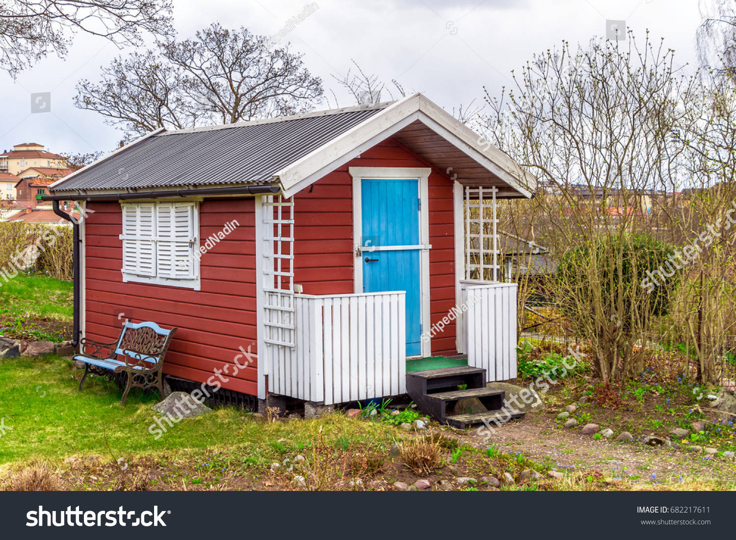 Red tiny house or shed at a plot of an allotment. Early spring time. Small red shack with blue door, white frames, shutters and metal forged bench by wall. Hut painted in traditional Swedish red color #682217611