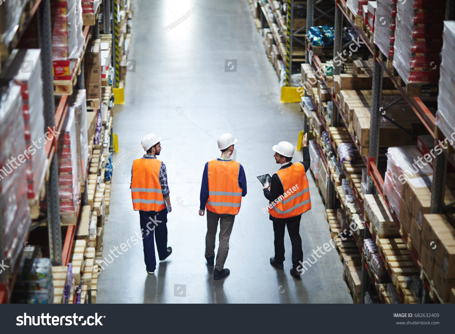 Group of warehouse workers wearing hardhats and reflective jackets waking in aisle between tall racks with packed goods, back view #682632409