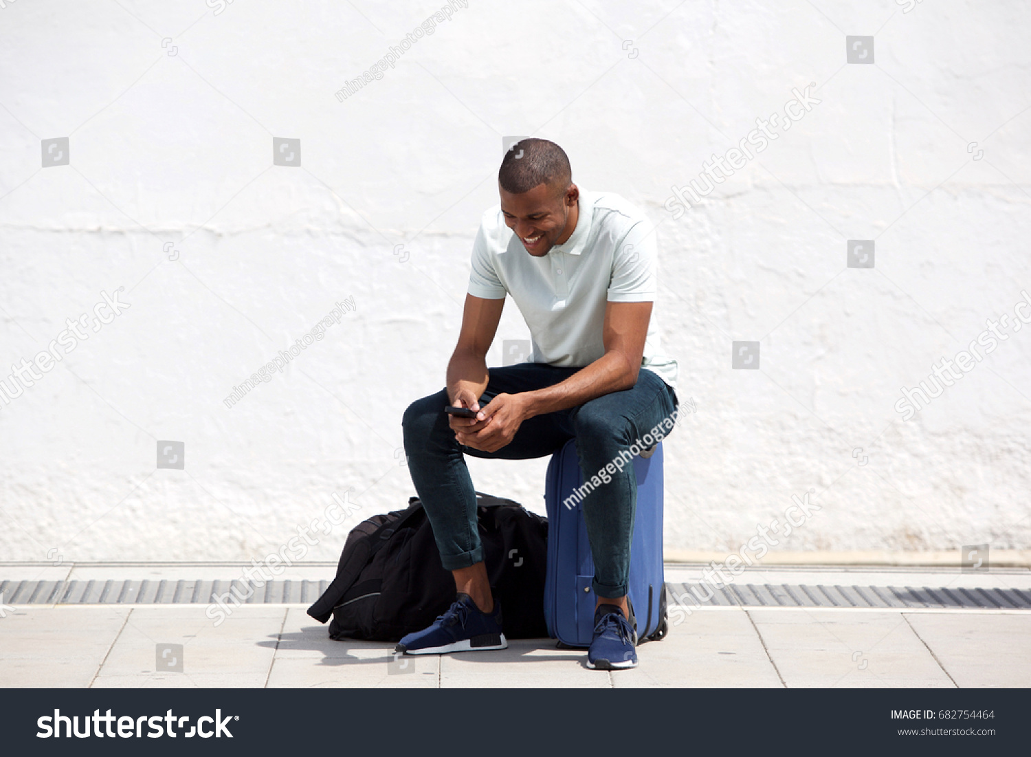 Full length portrait of african man sitting on suitcase and looking at mobile phone #682754464
