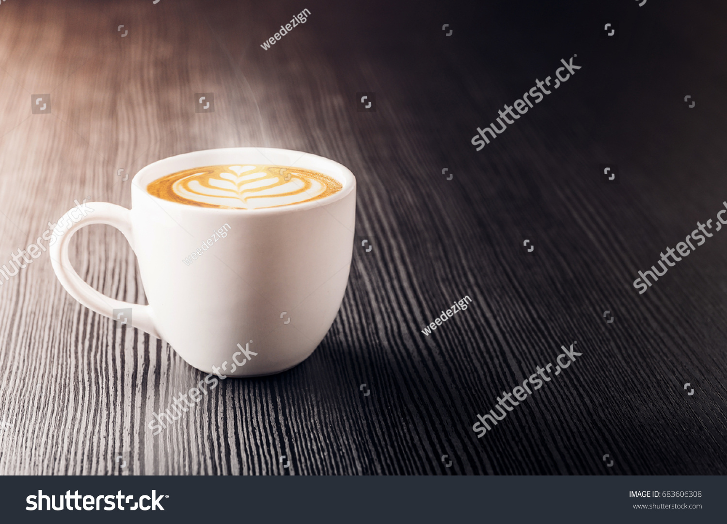 Close up white coffee cup with heart shape latte art foam on black wood table near window with light shade on tabletop at cafe. #683606308