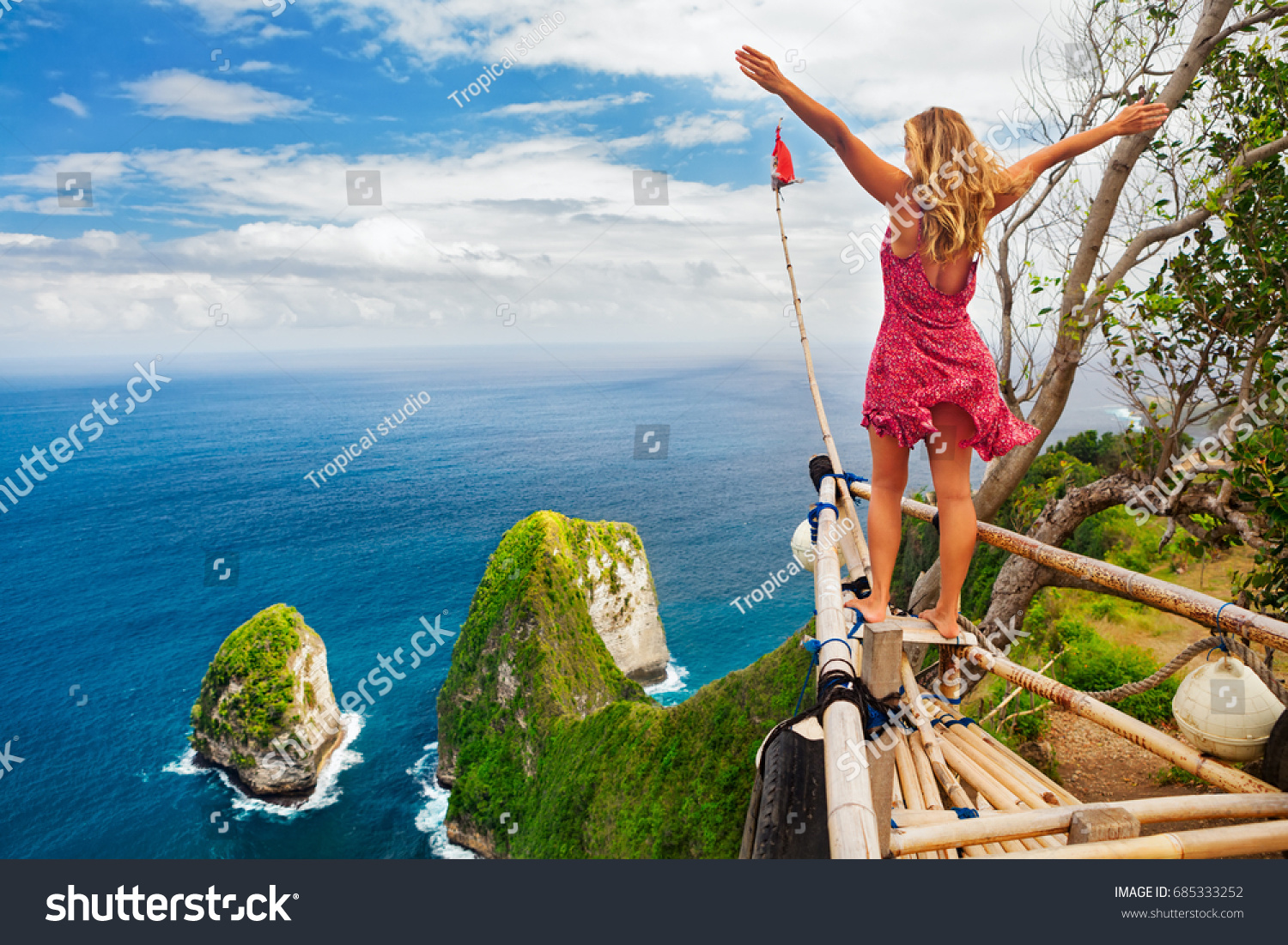 Family vacation lifestyle. Happy woman with raised in air hand stand at viewpoint. Look at Kelingking beach under high cliff. Travel destination in Bali. Popular place to visit on Nusa Penida island. #685333252