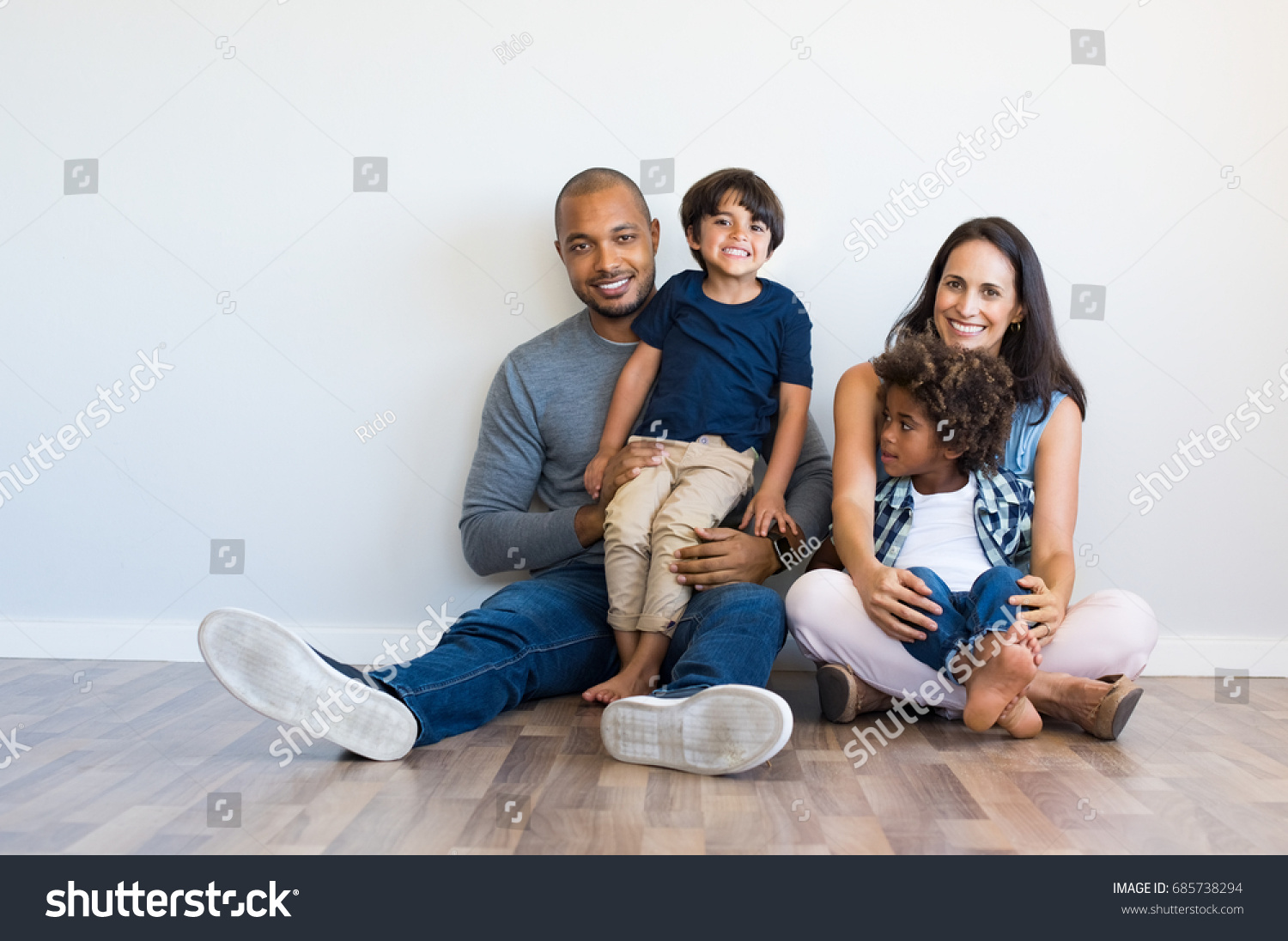 Happy multiethnic family sitting on floor with children. Smiling couple sitting with two sons and looking at camera. Mother and black father with their children leaning on wall with copy space. #685738294