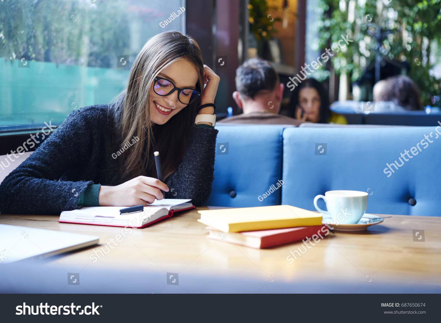 Positive female student in stylish eyeglasses with good lense writing down funny story in notebook sitting in coffee shop with cup of cappuccino and books.Smiling female noting text in notepad #687650674
