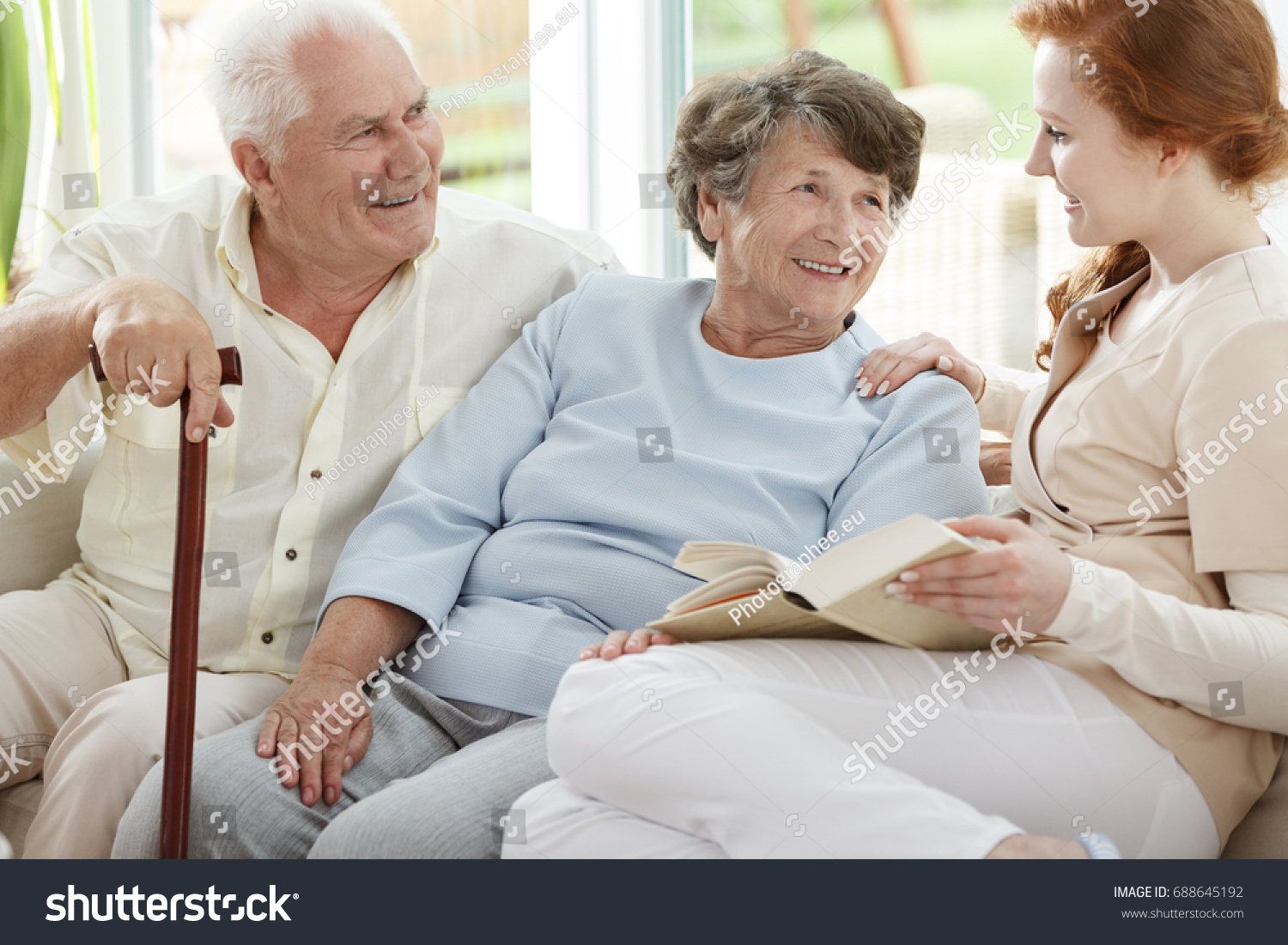 Two elder people are looking at smiling nurse who reads book in common room #688645192