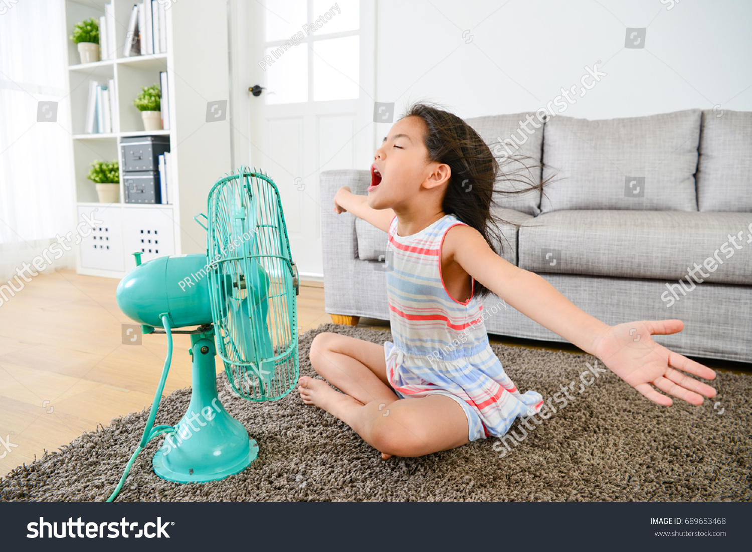 cheerful beauty girl kid opening arms singing enjoying cool wind when she sitting on living room floor blowing electric fan. #689653468