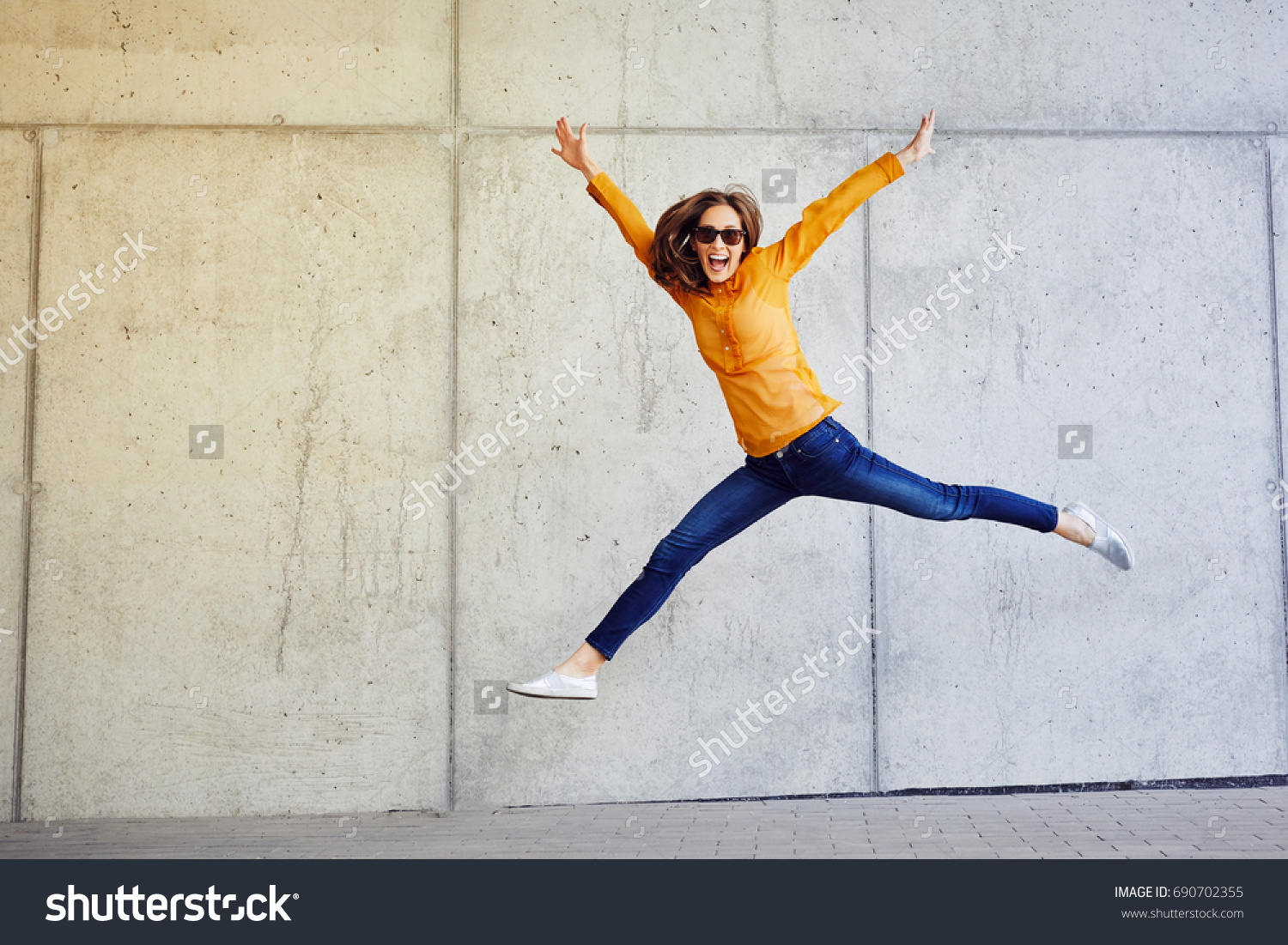 Joyful young lady jumping and raising arms in front of wall outside #690702355