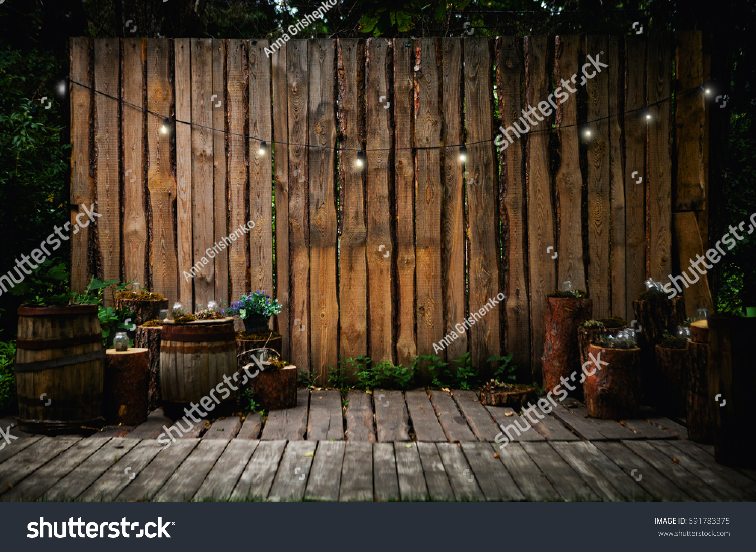 A beautiful recreation area in the park or in the garden. Wooden terrace, rustic loft interior. Retro garland with luminous bulbs, decorated pots with flowers and candles #691783375