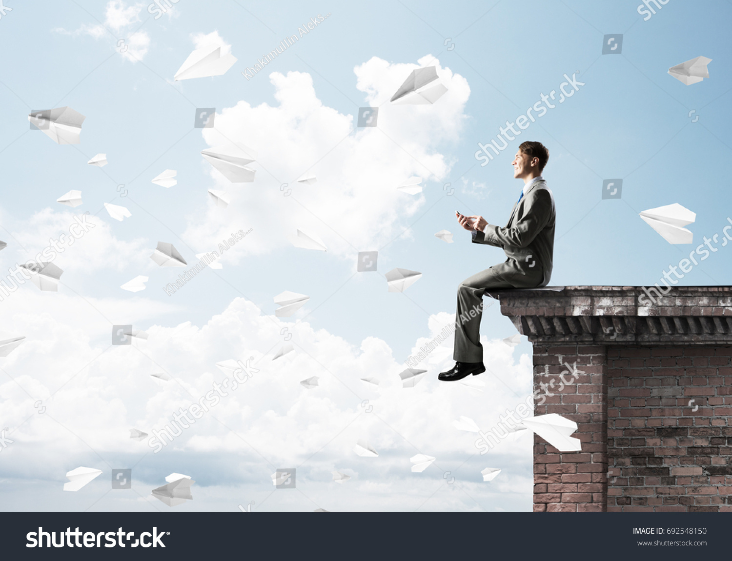 Young businessman sitting on building top with smartphone in hands #692548150