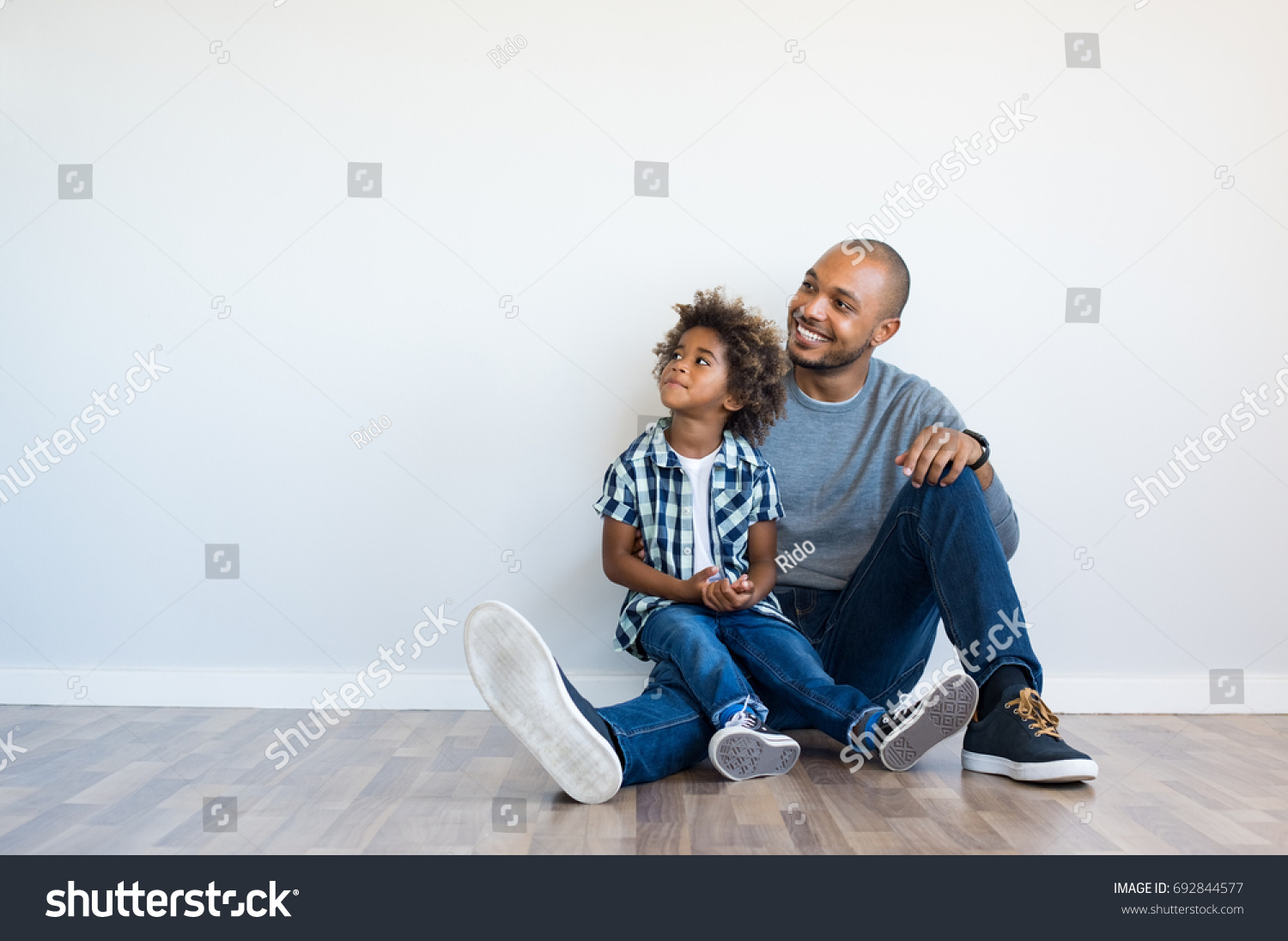 African father and his son sitting on floor and looking up in a blank wall. Happy dad and little boy sitting in an empty room. Young black man with his child thinking and pensive with copy space. #692844577