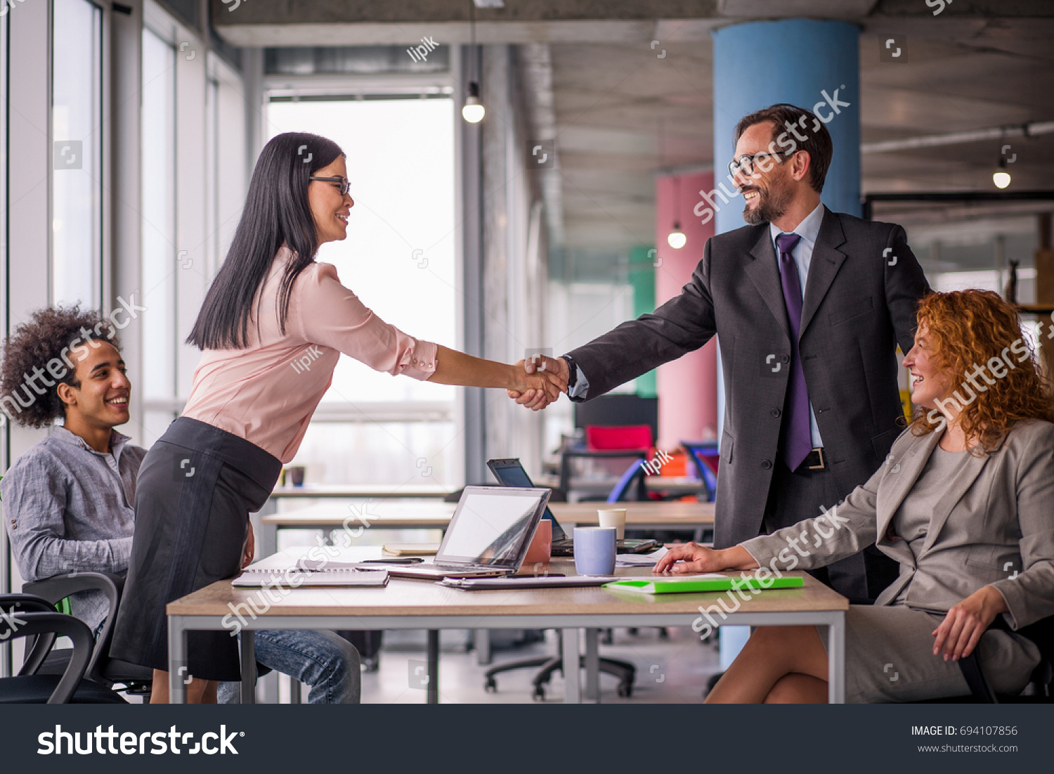 Two business teams successfully negotiating, shaking hands. At meeting table business groups shaking hands on completed deal. Man and woman handshake. #694107856