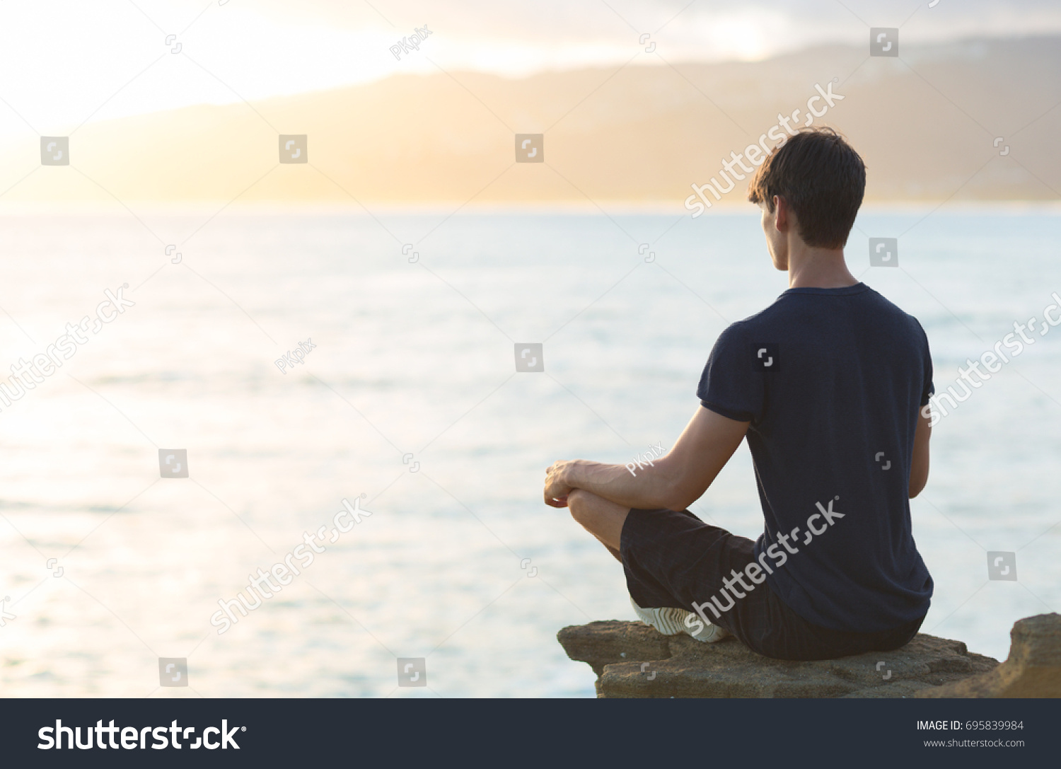 Young man meditating on top ocean cliff during sunset.  #695839984