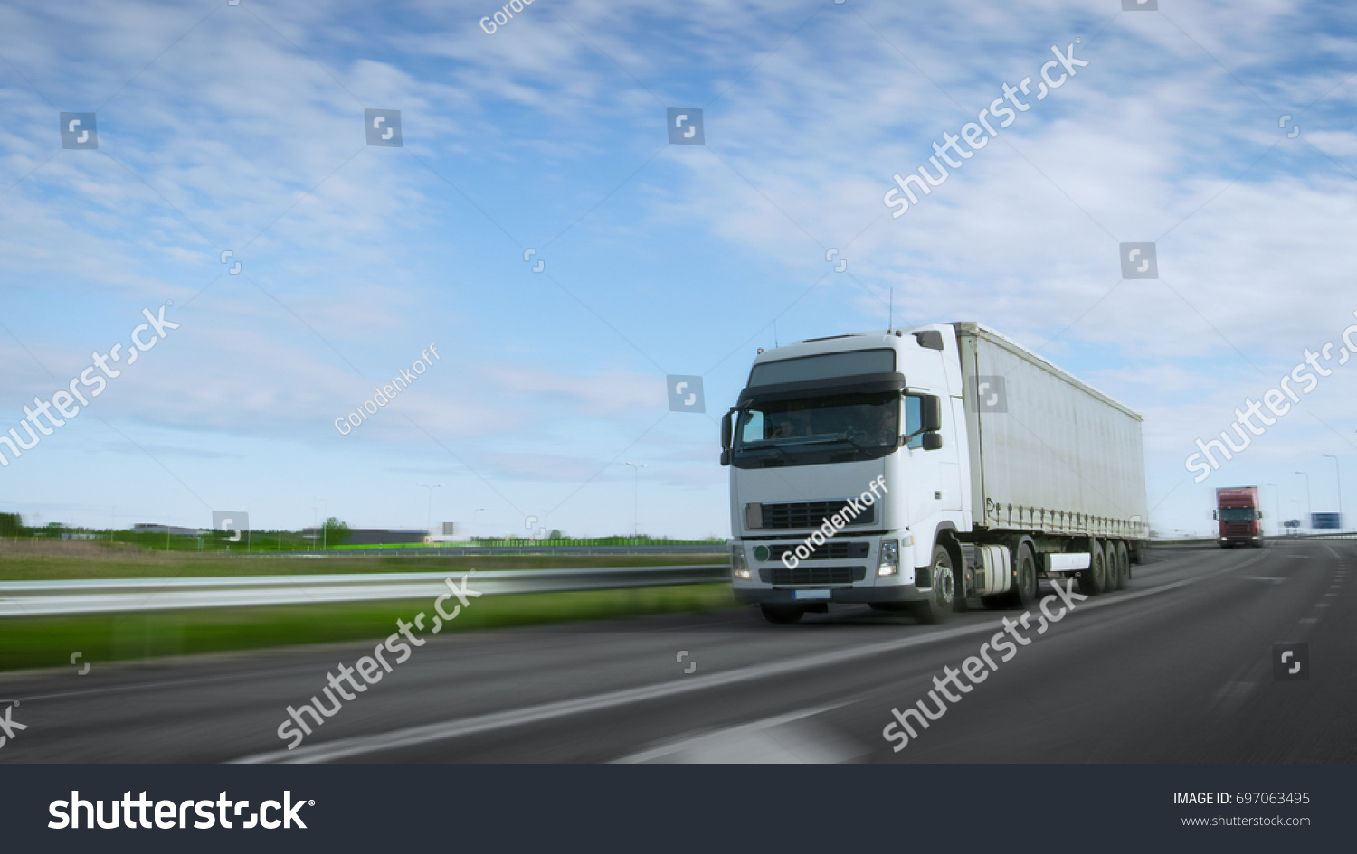 Speeding White Semi Truck with Cargo Trailer Drives on the Highway. Truck is First in the Column of Heavy Vehicles, Sun is Shining. Blur motion. #697063495