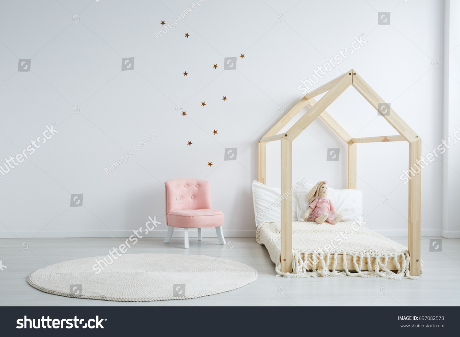 Modern children's furniture in a spacious bedroom with star stickers on the white wall, and a pastel pink comfortable chic chair next to a wooden bed #697082578