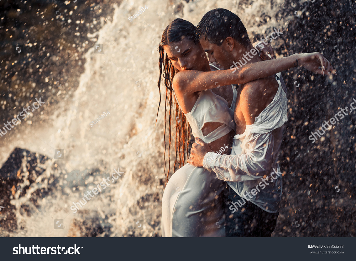 Young enamored wet couple hugs under spray and drops of waterfall. Around them are visible jets and streams of running water.  #698353288