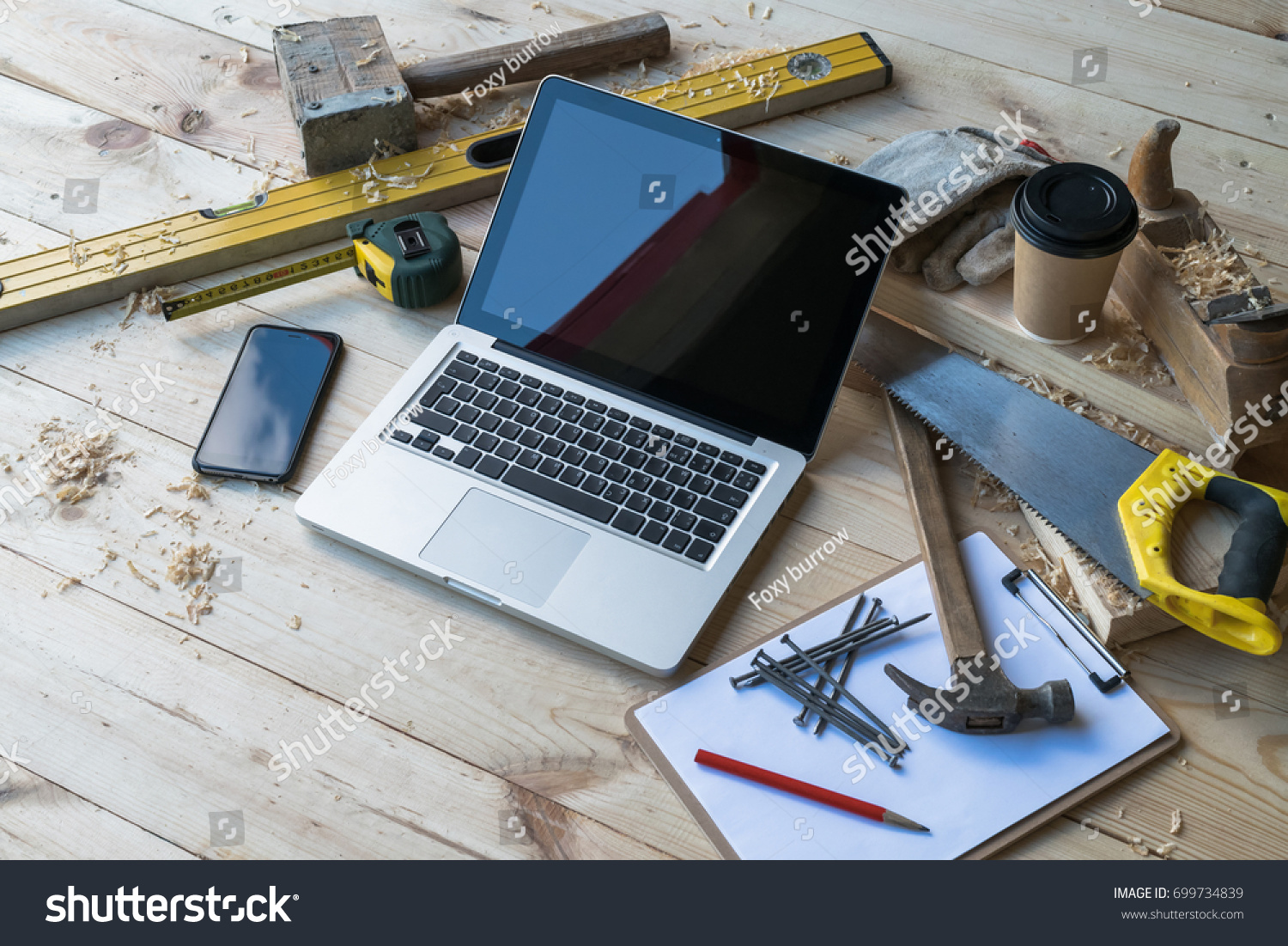 Close up laptop on wooden planks, smartphone, clipboard, pen, nails, hammer, plane, saw, tape measure, level, gloves, glasses, cup of coffee, sawdust. Carpentry. Mock up. Construction business. #699734839