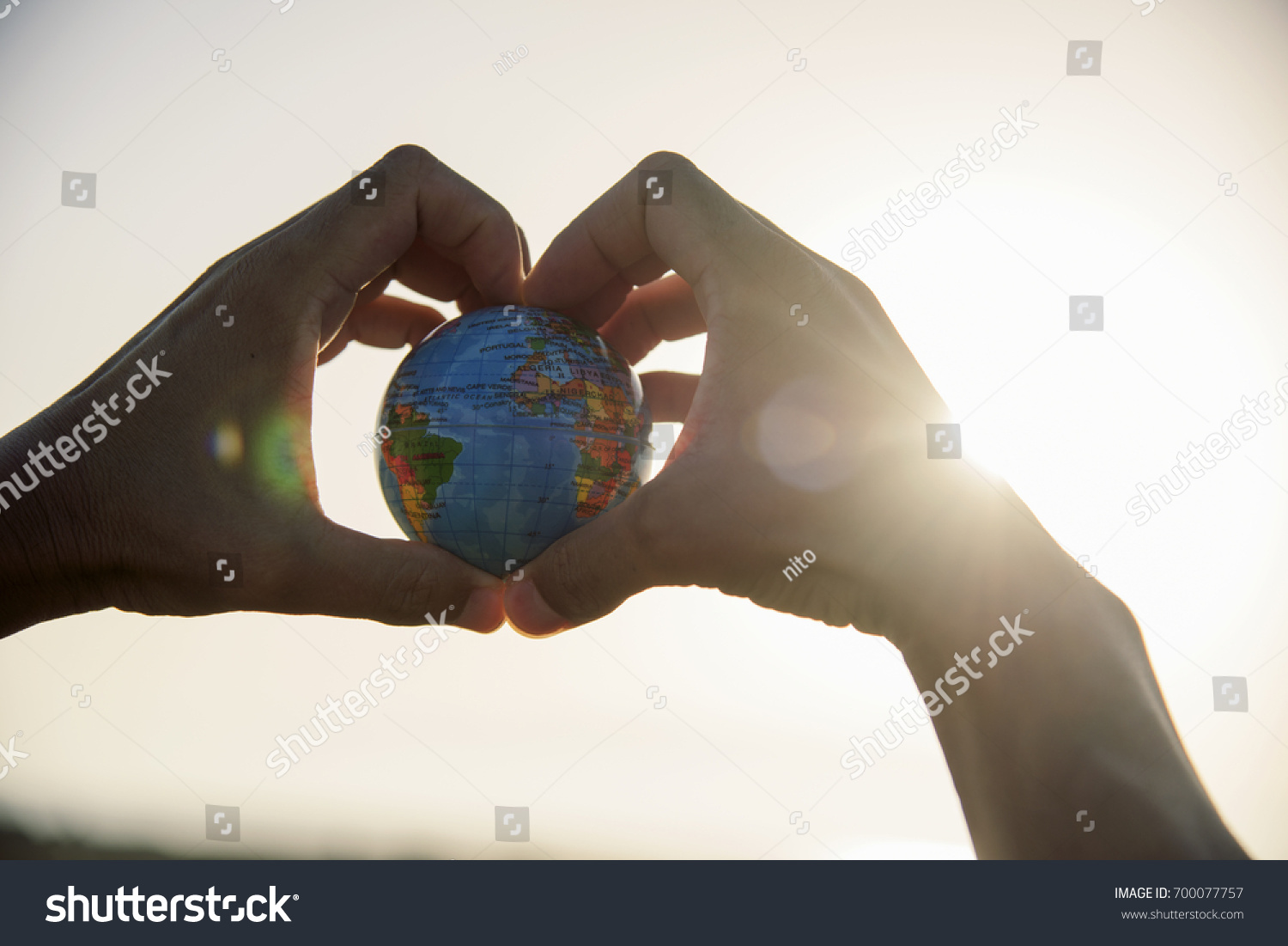 closeup of a young caucasian man with a world globe in his hands forming a heart against the sky and a ray of sun showing up on the right #700077757