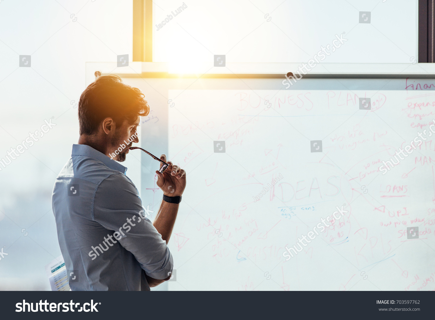 Business investor in deep thought looking at the business ideas written on the whiteboard. Businessman thinking while holding his spectacles to mouth #703597762