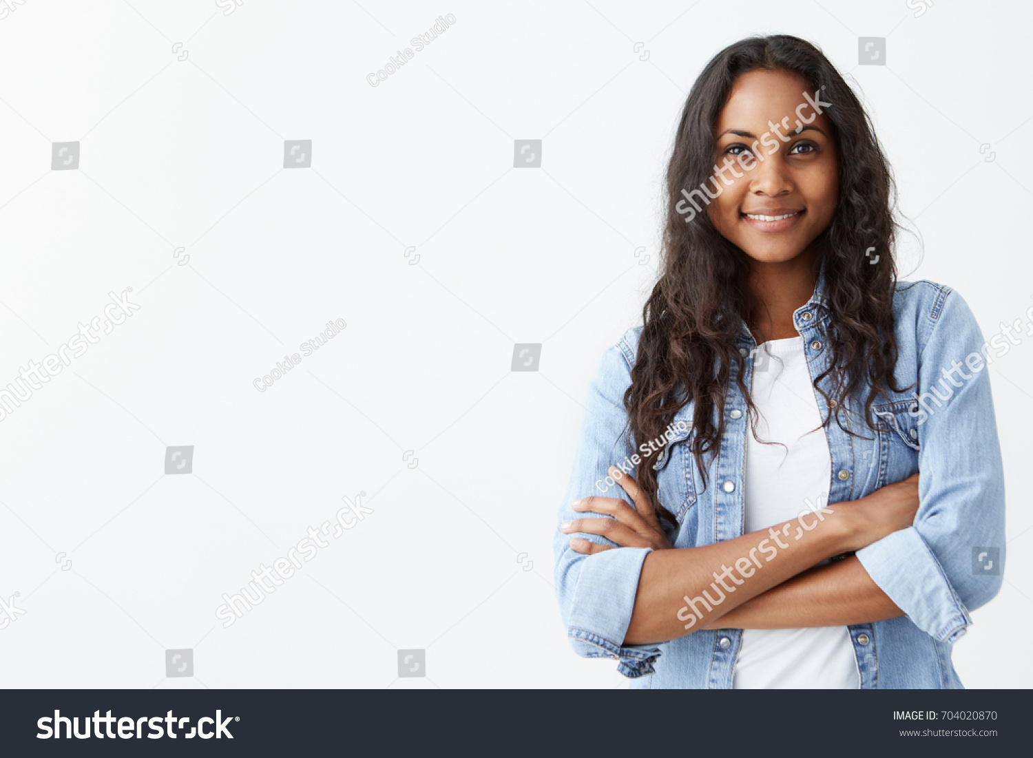 Glad young good looking Afro-American woman with clean dark skin and black long hair posing indoors with crossed arms, smiling broadly with white teeth, laughing at good joke, wearing denim shirt over #704020870