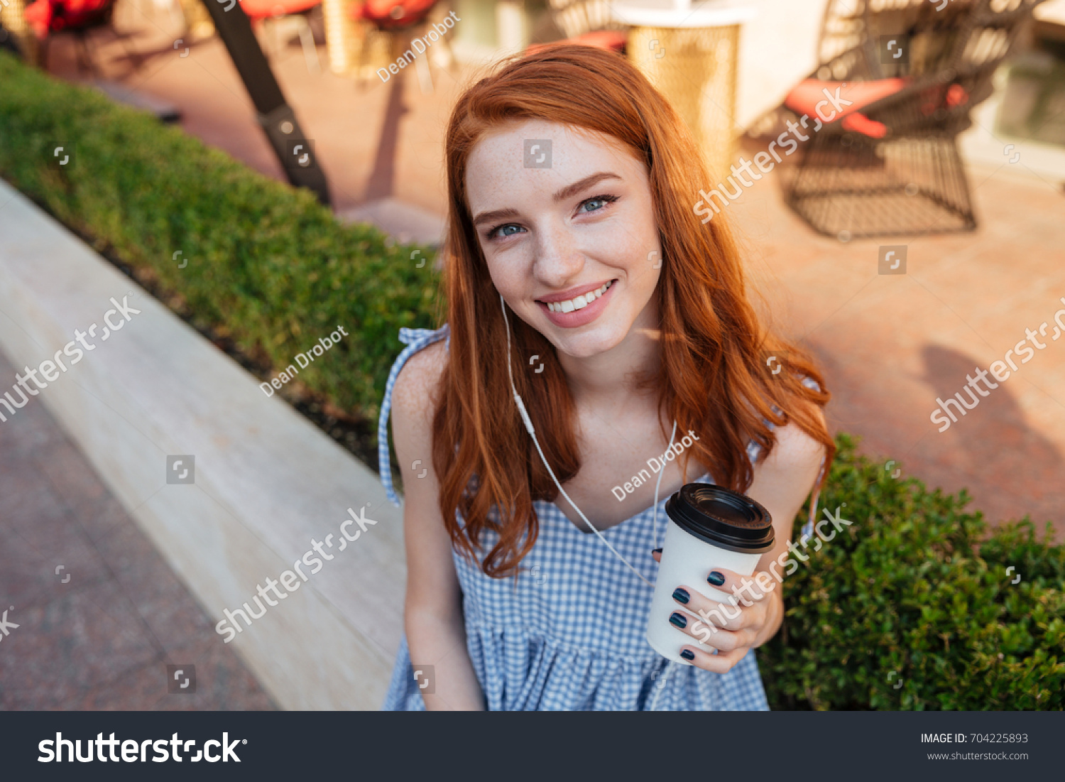 Close up of a young smiling redhead girl in earphones holding cup of coffee and looking at camera while sitting outdoors and listening to music #704225893