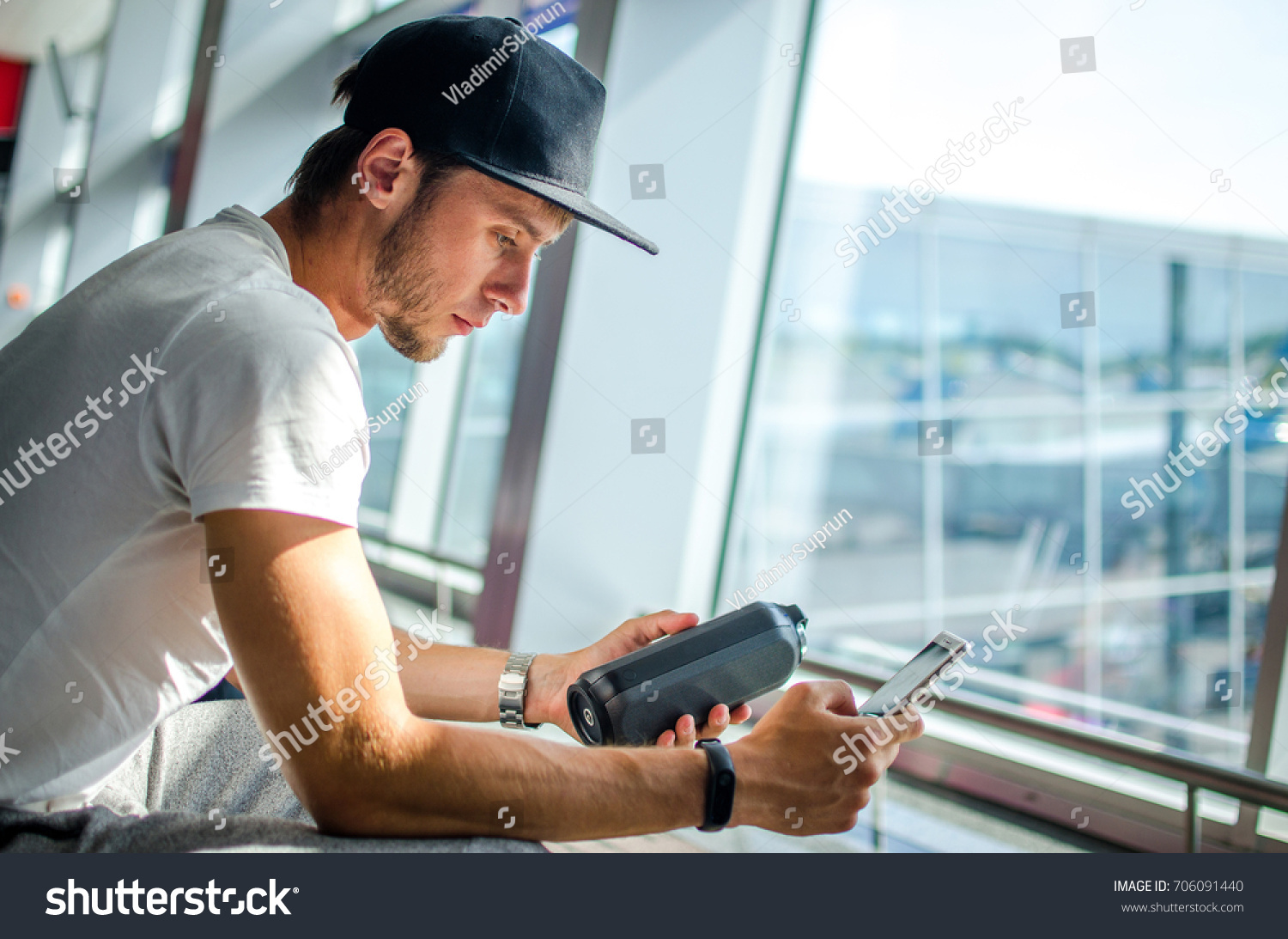 The guy sits at the airport terminal and listens to music on the portable speaker #706091440