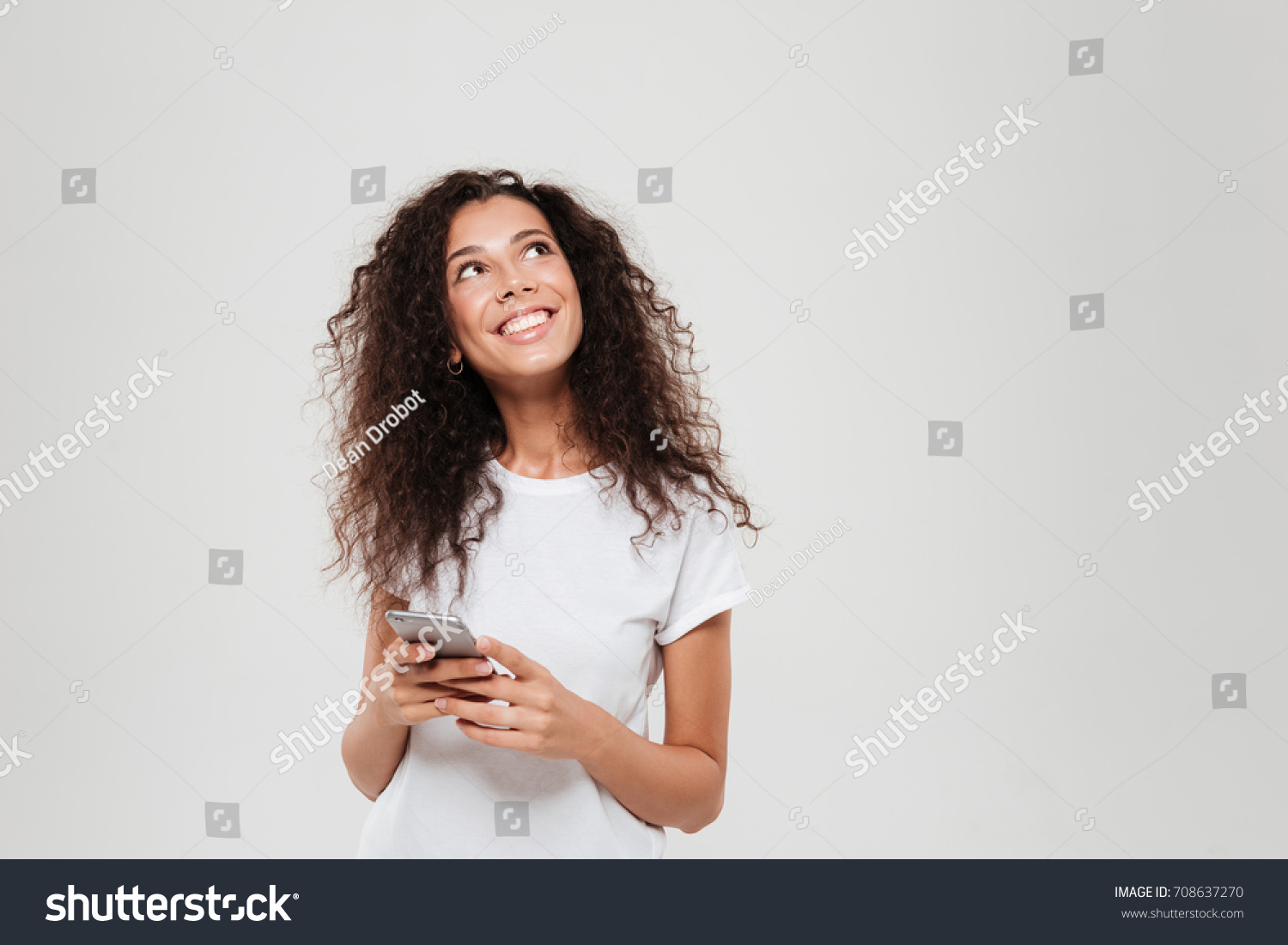 Smiling pensive woman holding smartphone in hands and looking up over gray background #708637270