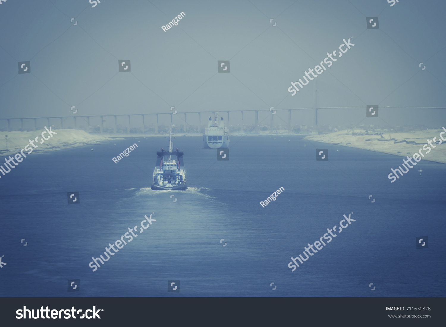 In the Suez Canal - a military ship and a tugboat forward to the city of  El Qantara and the Mubarak Peace Bridge in foggy evening mood #711630826