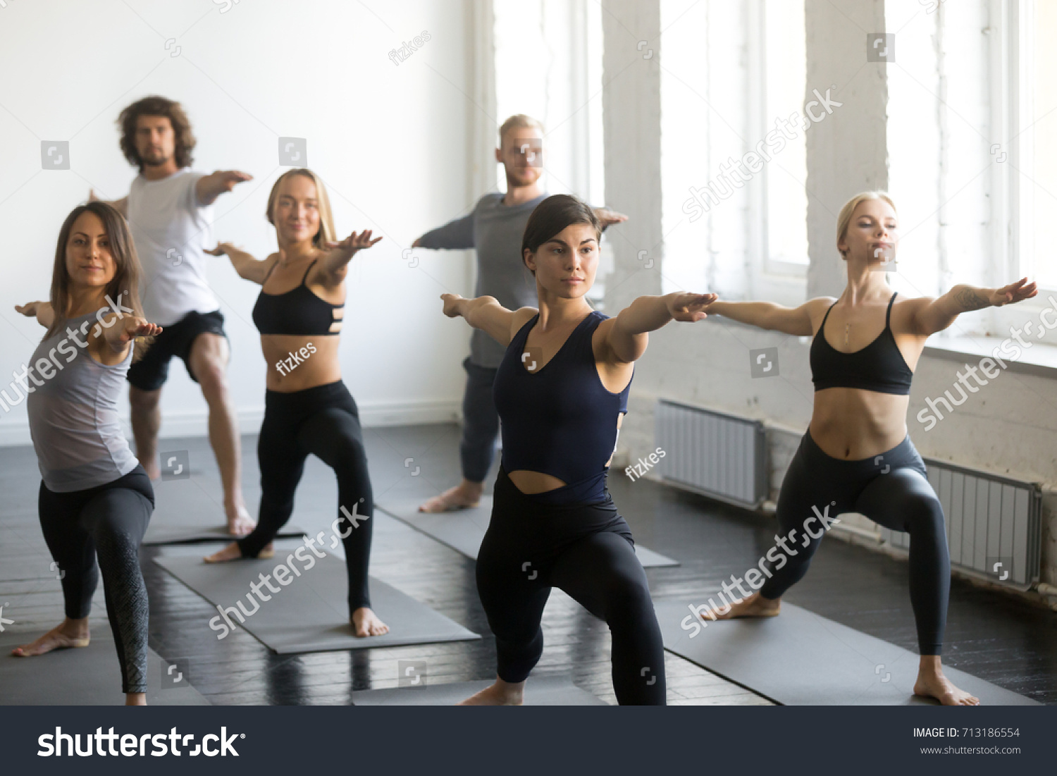 Group of young sporty attractive people practicing yoga lesson with instructor, standing in Warrior Two exercise, Virabhadrasana pose, working out, indoor, studio. Wellbeing, wellness concept  #713186554