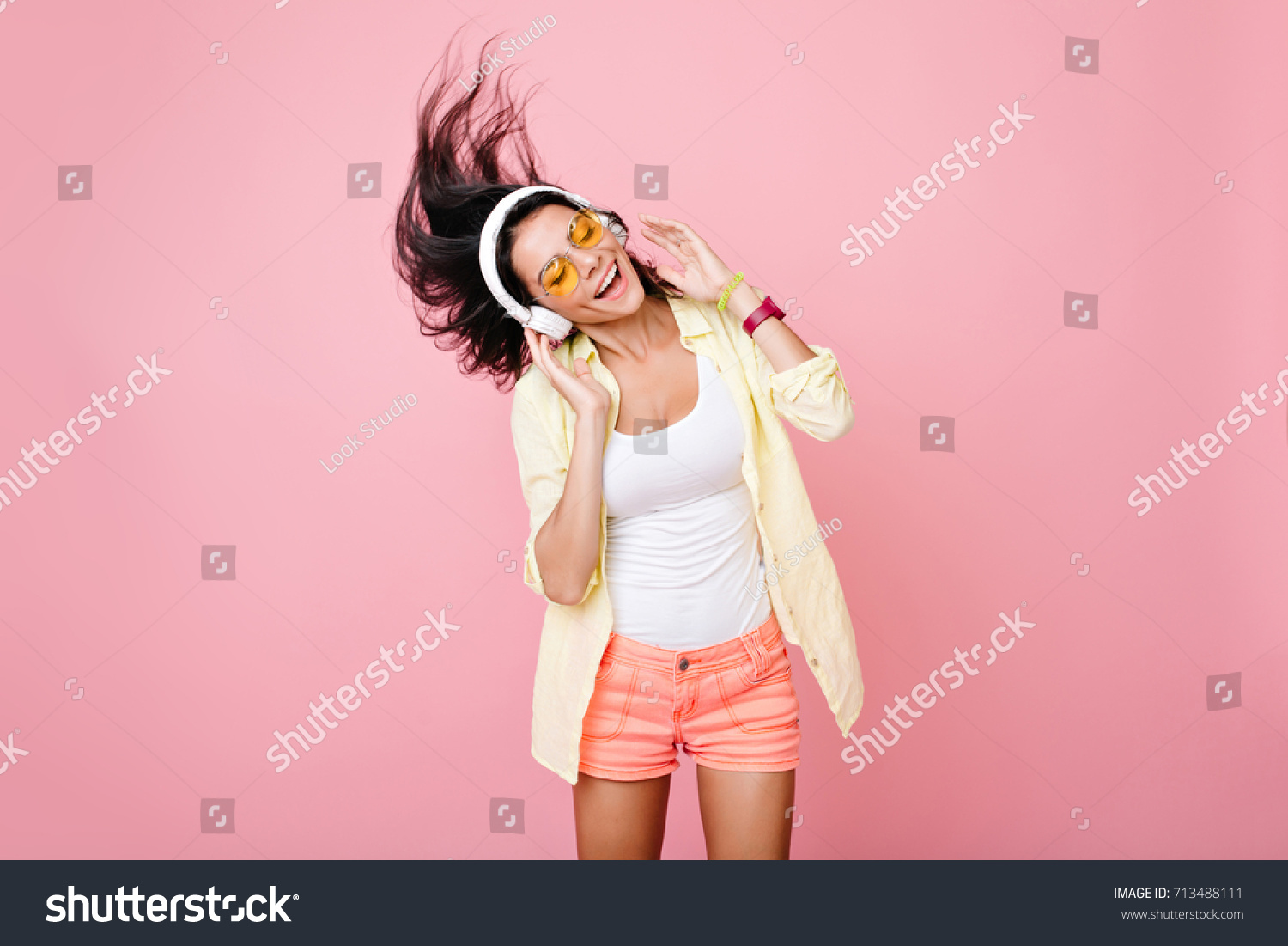 Gorgeous brunette lady in yellow glasses listening music in headphones and singing on pink background. Charming girl in shorts and earphones dancing with hair waving and eyes closed. #713488111