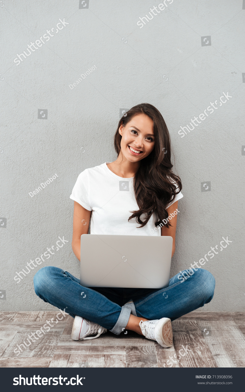 Happy young asian woman working on laptop computer while sitting on the floor with legs crossed isolated over gray background #713908096