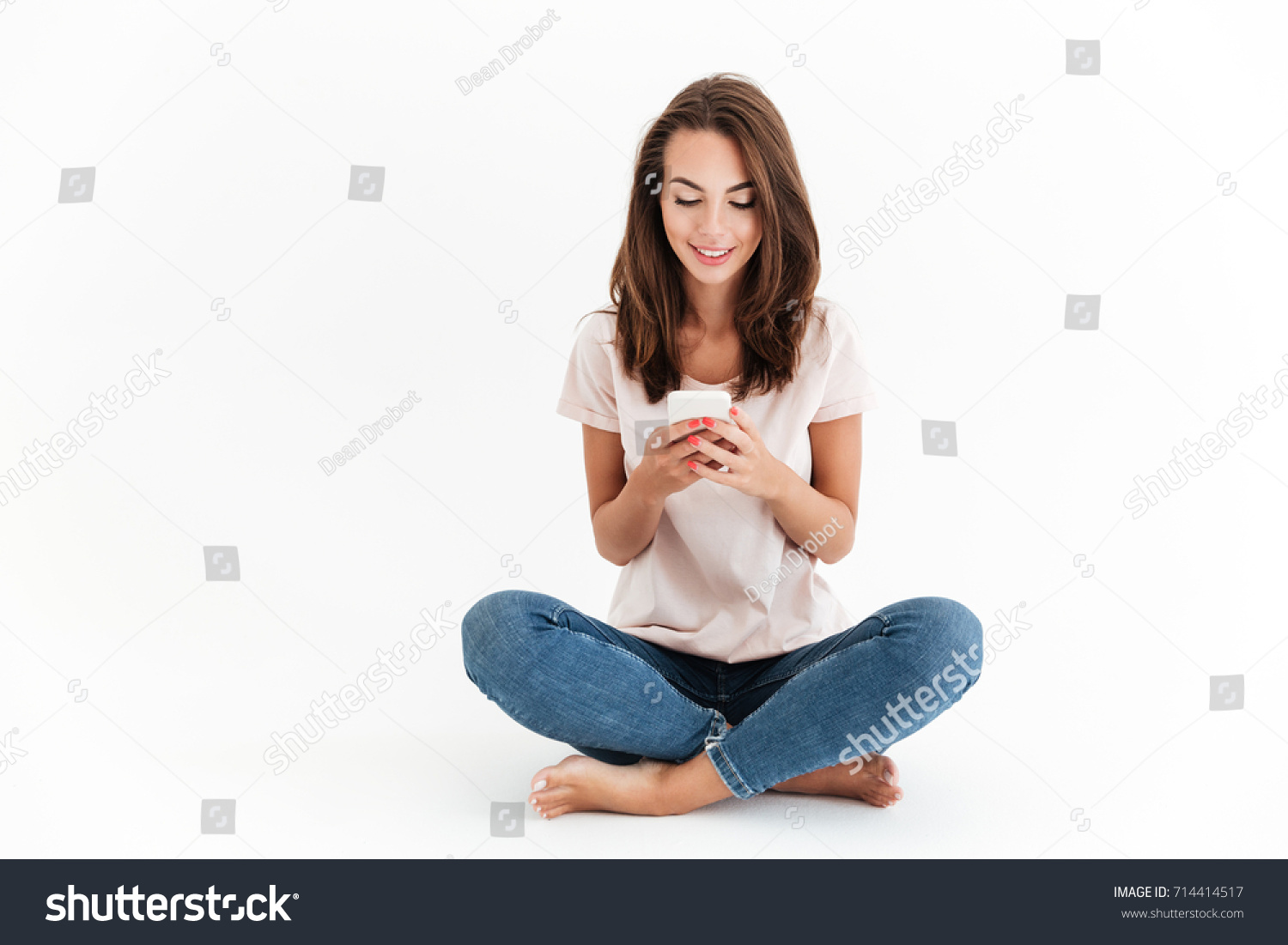 Smiling brunette woman sitting on the floor and using smartphone over white background #714414517