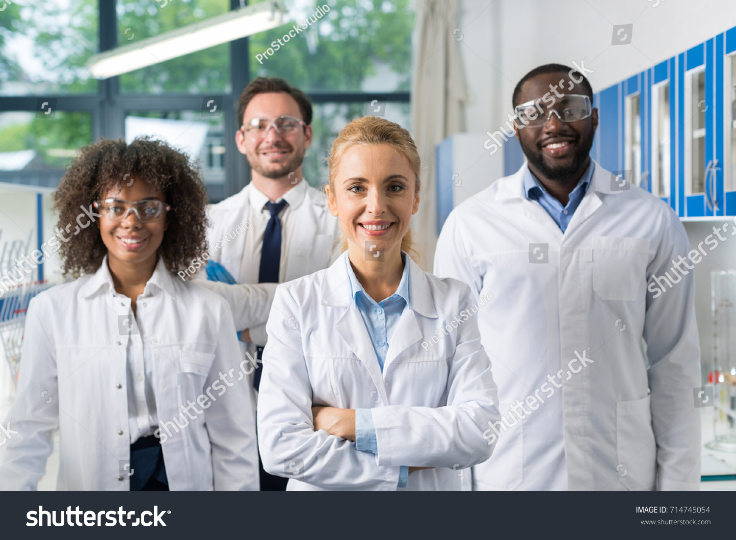 Smiling Group Of Scientists In Modern Laboratory With Female Leader, Mix Race Team Of Scientific Researchers In Lab Wearing White Coats And Protective Glasses #714745054