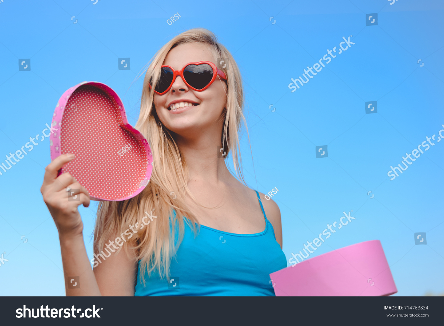 Joyful excited lovely woman holding heart shaped gift box in hands. Happy smiling surprised young lady over light blue sky background. Excitement beauty celebrating moment #714763834