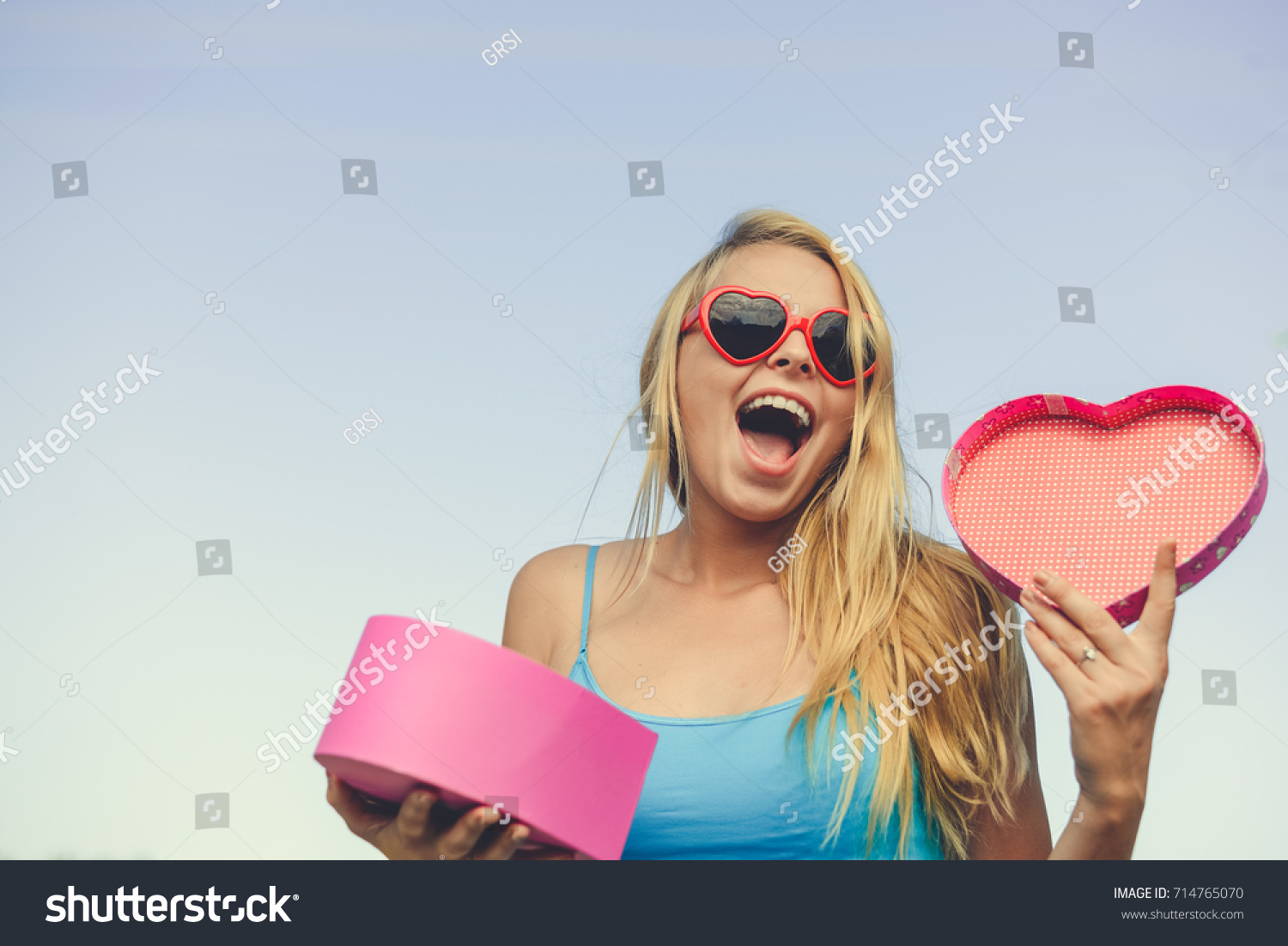 Joyful excited lovely woman holding heart shaped gift box in hands. Happy smiling surprised young lady over light blue sky background. Excitement beauty celebrating moment #714765070