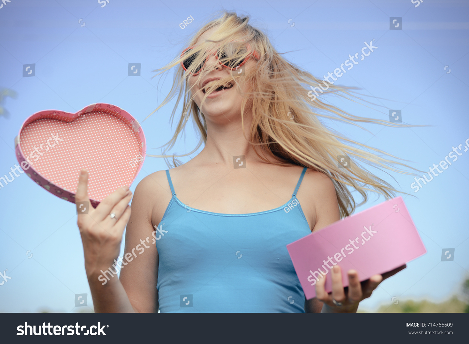 Joyful excited lovely woman holding heart shaped gift box in hands. Happy smiling surprised young lady over light blue sky background. Excitement beauty celebrating moment #714766609