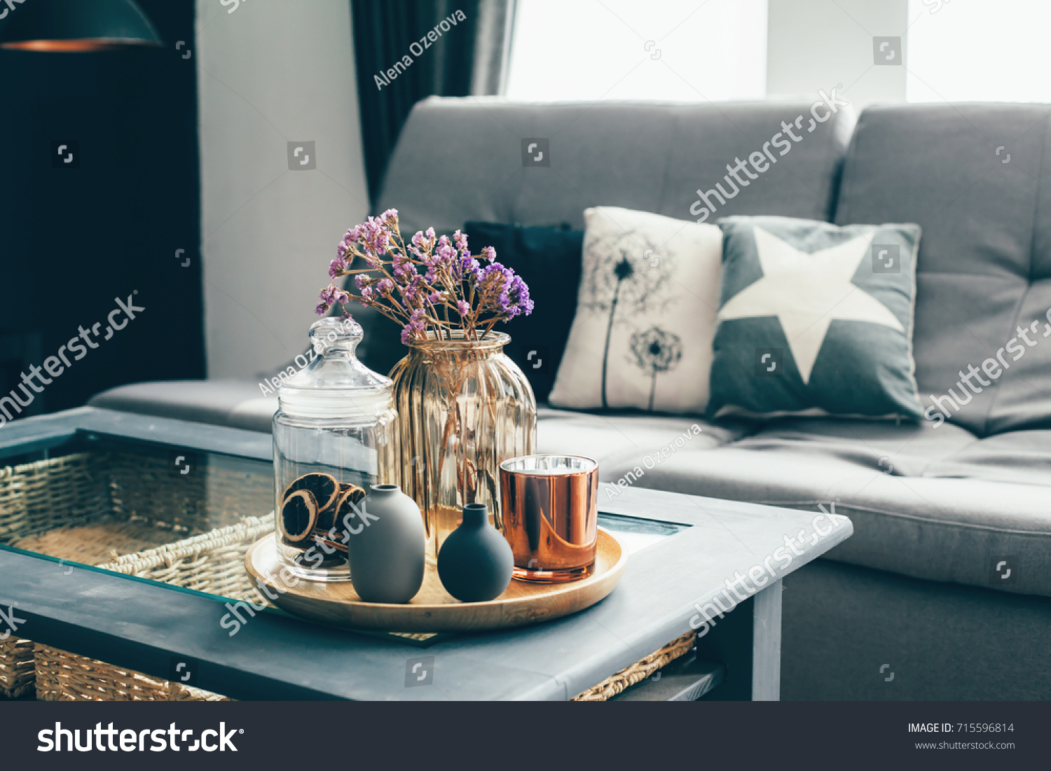 Home interior decor in gray and brown colors: glass jar with dried flowers, vase and candle on the wooden tray on the coffee table over sofa with cushions. Living room decoration. #715596814