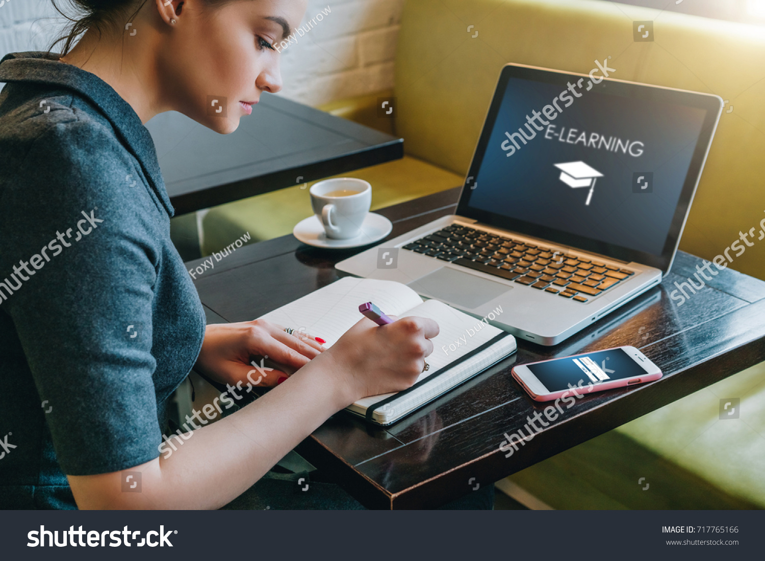 Young businesswoman sitting at table in cafe in front of laptop with inscription on screen e-learning and image of square academic cap and making notes in notebook,diary. Online education,e-learning. #717765166