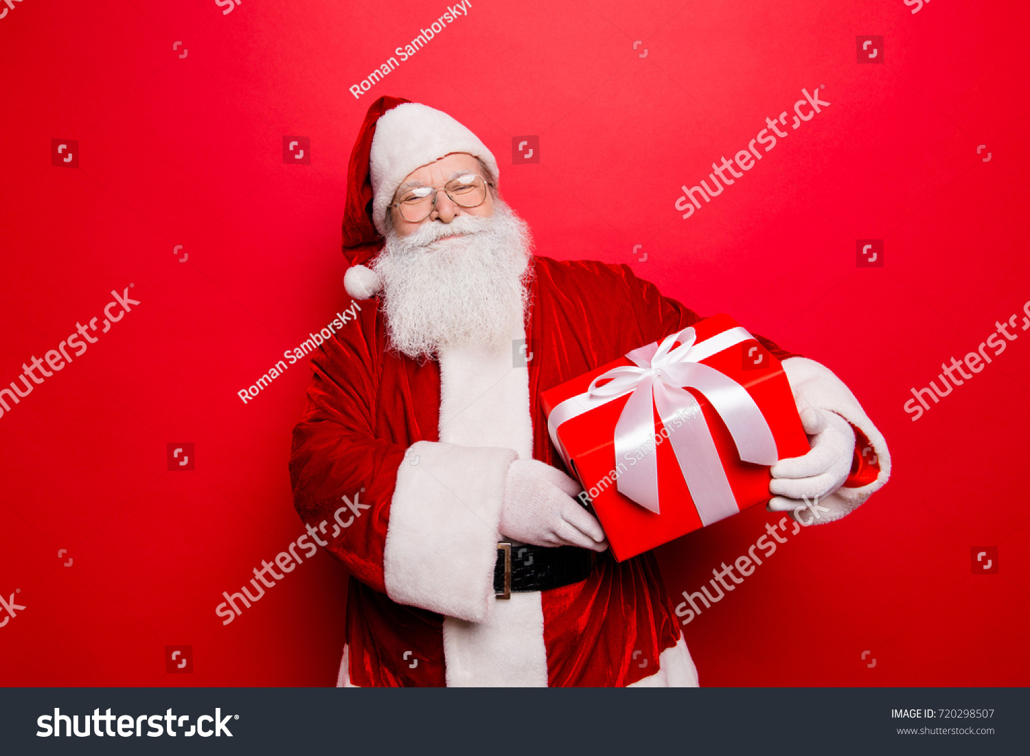 Happiness, noel, festive seosonal occasion. Funny Saint Nicholas in red traditional fur coat and head wear with a wrapped gift with bow, isolated on red background, wishing holly jolly x mas #720298507
