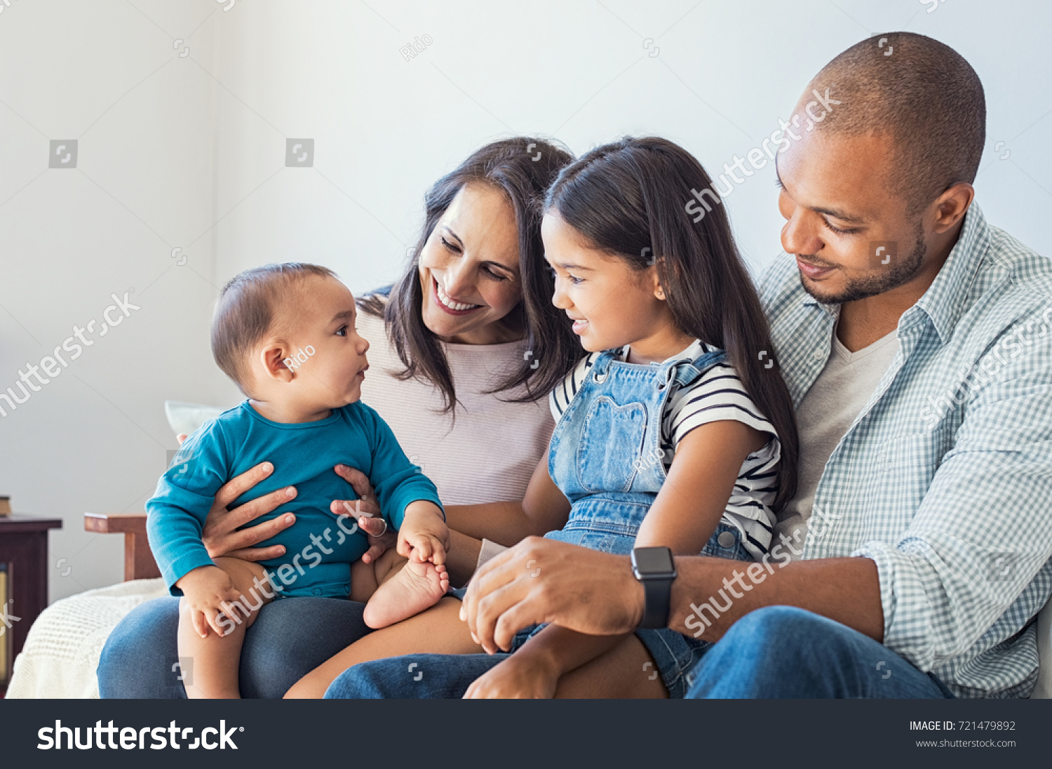 Multiethnic family playing with happy baby son at home. Parent and children relaxing together on the sofa at home in the living room. Little girl sitting on leg of dad looking her new cute brother. #721479892