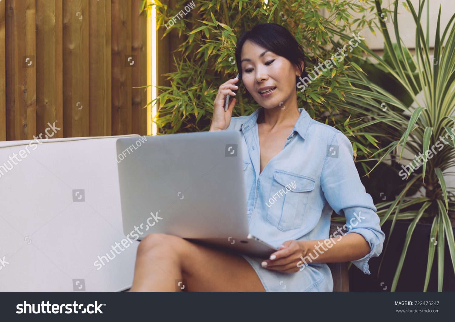Young business woman is having call while sitting in a modern coffee shop with a portable computer on her knees. Asian female is shopping online by a smartphone and a laptop connected to the internet. #722475247