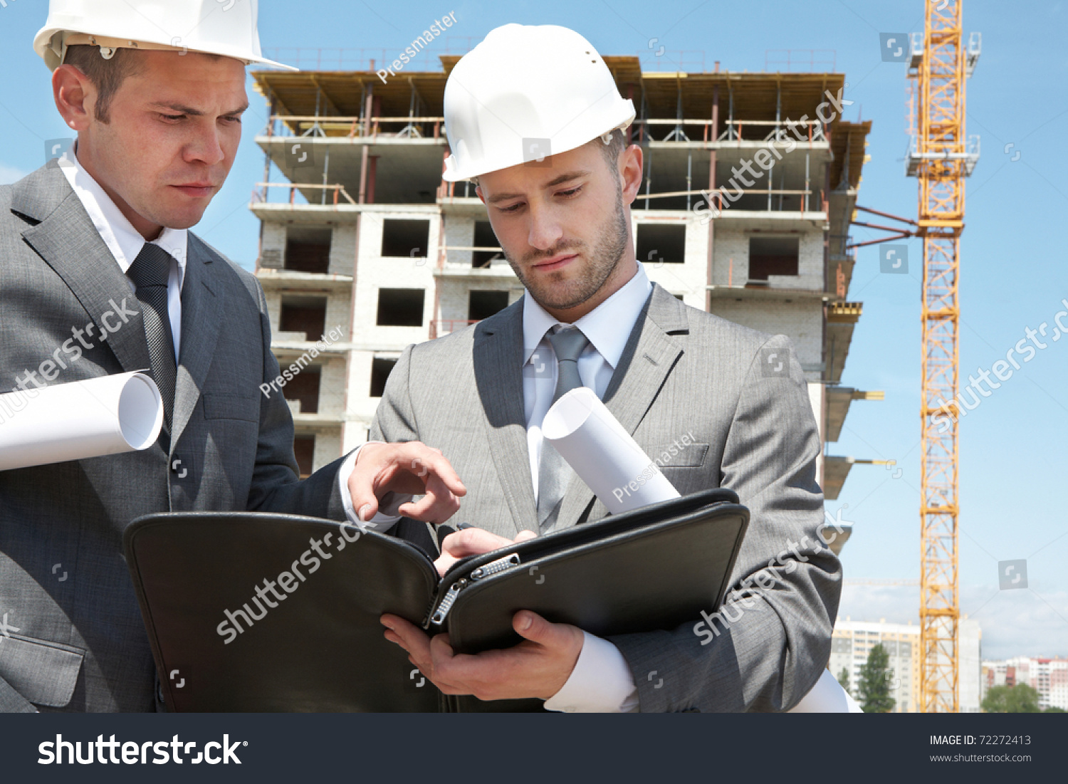 Portrait of two builders standing at building site and discussing new project held by one of them #72272413
