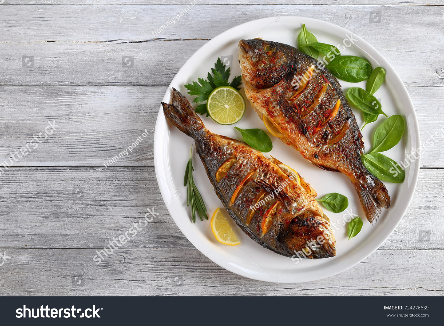 delicious grilled dorado or sea bream fish with lemon and orange slices, spices, fresh parsley and spinach on white platter on old wooden table, horizontal view from above #724276639