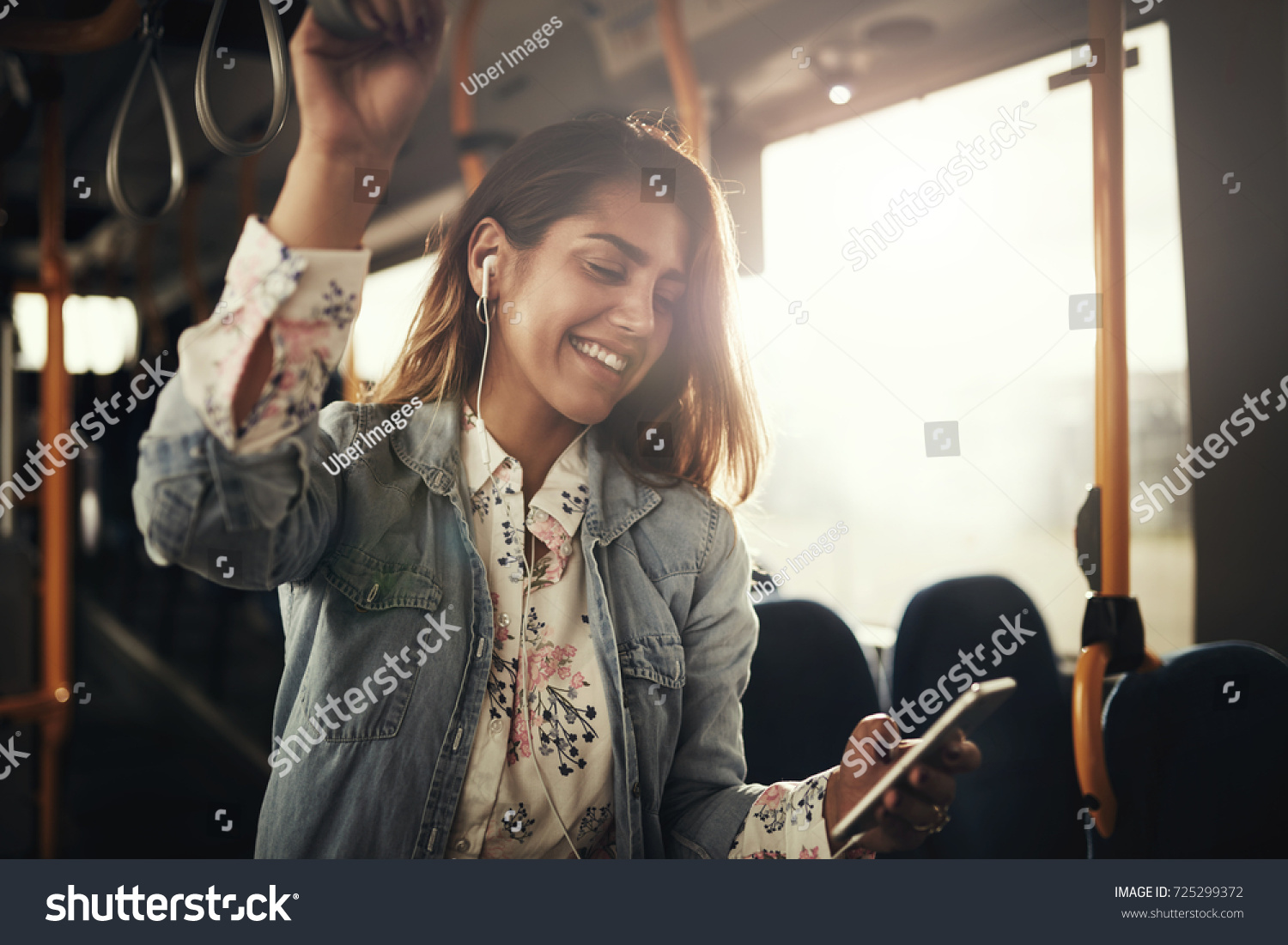 Young woman smiling while standing by herself on a bus listening to music on a smartphone  #725299372