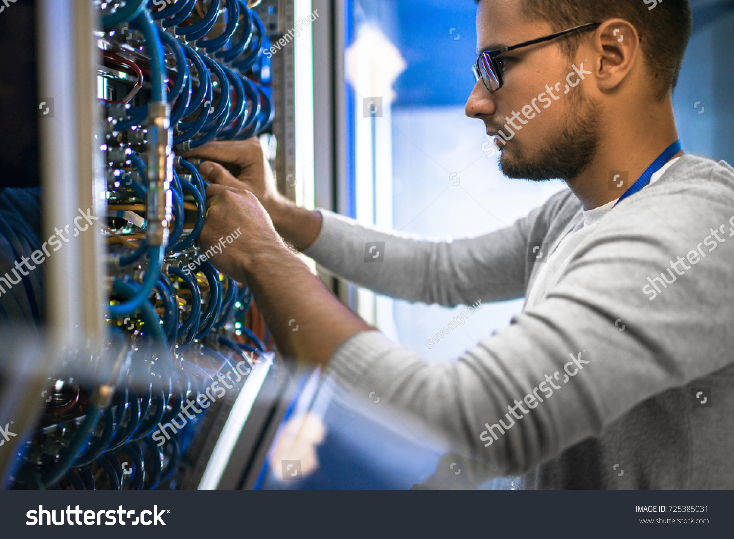 Side view portrait of young man wearing glasses connecting cables in server cabinet while working with supercomputer in data center #725385031